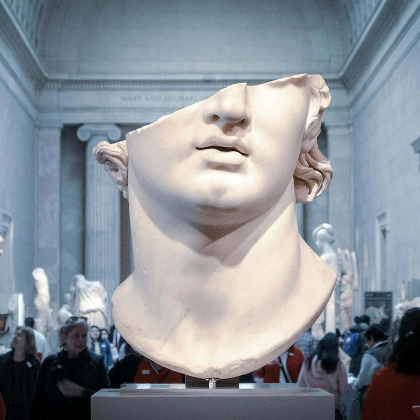 A large, fragmented marble head from an ancient classical sculpture displayed on a pedestal in a crowded museum gallery.